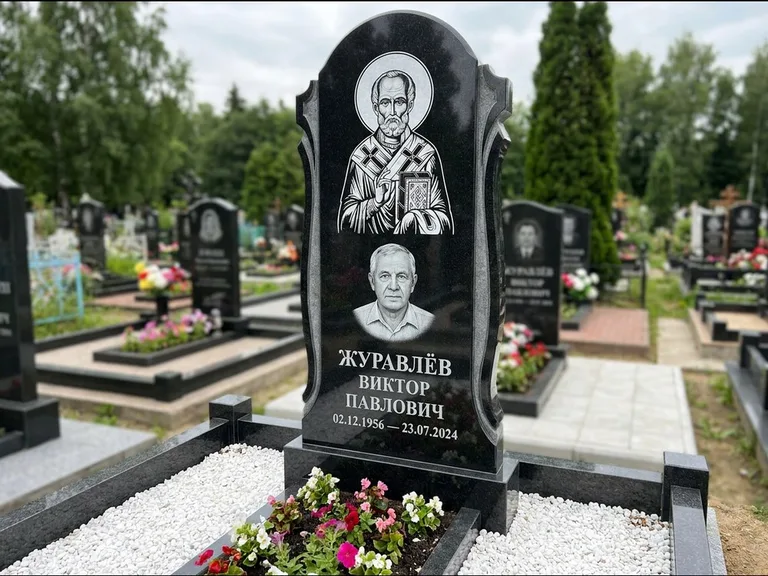 Памятник с гравировкой иконы: стела с образом в полный рост на обороте — гравировка образа Матроны Московской, низкие кусты спиреи в цвету, работа «Лебедева» в Красноярске